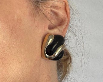 May include: Square-shaped black and gold earrings. The earrings have a gold-colored frame with a black center and a gold-colored, curved design. The earrings are a bold, geometric statement piece.