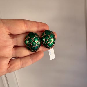 Puede incluir: Un par de pendientes ovalados de clip. Los pendientes presentan un diseño de esmalte verde oscuro con un contorno de metal dorado. Los pendientes tienen un patrón texturizado y orgánico.