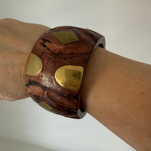 Può includere: Un bracciale largo in legno con finitura marrone rossiccio e inserti in ottone. Il bracciale presenta forme geometriche, tra cui quadrati e semicerchi. Il bracciale è largo circa 6,35 cm, aggiungendo un tocco di eleganza al design.
