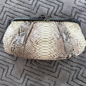 Pode incluir: Uma clutch com um padrão de pele de cobra em tons de creme, cinza e castanho. A bolsa tem uma moldura preta e um fecho decorativo. A textura da pele de cobra é visível.