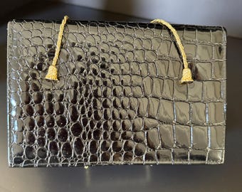 May include: Black rectangular clutch with a glossy, crocodile-skin texture. The clutch features two gold-colored, beaded tassels attached to the top. The bag is a classic accessory for formal events.