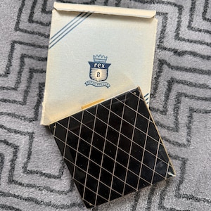 Op de afbeelding: Een vintage zwarte compactpoederdoos met een gouden sluiting en een ruitpatroon. De compactpoederdoos is open en onthult een spiegel. De doos is lichtblauw met de tekst "rex" en een wapen. De compactpoederdoos ligt op een grijs patroon.