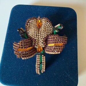 May include: A detailed brooch in the shape of a flower, featuring a central cluster of clear rhinestones and petals in shades of purple and gold. Accents include green and orange gemstones. The brooch is set on a blue velvet surface.