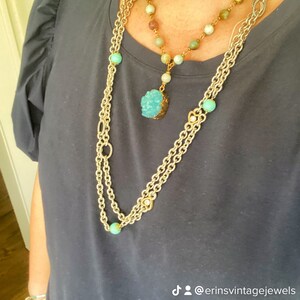 Könnte beinhalten: Ein mehrschichtiges Halsketten-Set mit einer langen Goldkette mit türkisfarbenen Perlen und einer kürzeren Halskette mit bunten Perlen. Der Fokus liegt auf einem Anhänger mit einem blauen Kristallcluster. Der Schmuck wird auf einem dunkelblauen Oberteil präsentiert.