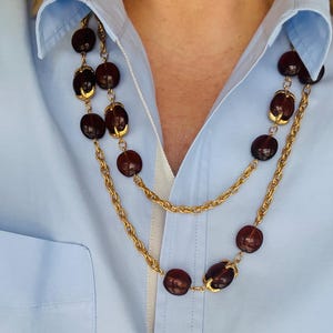 May include: A layered necklace with large, dark brown beads and gold-coloured accents. The beads are strung on a delicate gold-toned chain. The necklace is displayed against a light blue collared shirt.