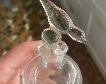 May include: A clear glass perfume bottle with a round body and a stopper featuring a bird design. The bird is perched atop the stopper, adding a decorative touch to the bottle. The bottle is held in a hand.
