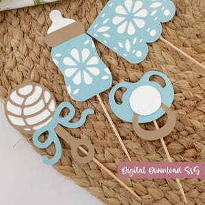May include: A set of four light blue and tan paper cutouts on wooden sticks, featuring a baby bottle, a pacifier, a rattle, and a decorative shape, all with intricate white floral-inspired cutouts and the text Digital Download SVG.