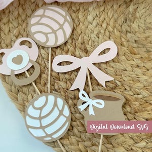 May include: A set of five paper cupcake toppers on wooden sticks featuring a pacifier, a bow, a mug with a small bow, and two concha sweet bread shapes in shades of light pink, white, and tan. The image includes the text Digital Download SVG.