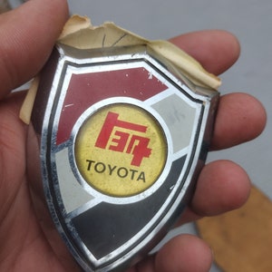 May include: A silver and black shield-shaped emblem with a red, white, and yellow circle in the center. The circle contains the text "TOYOTA" and a stylized red and white logo.