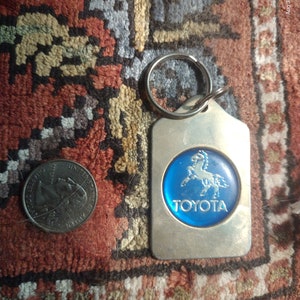 May include: A silver keychain with a blue and white Toyota logo featuring a horse. The logo is surrounded by a silver ring.