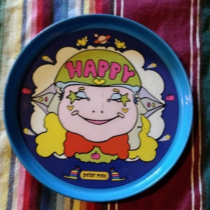 May include: A blue plastic tray with a colorful illustration of a smiling face wearing a green hat with the word "HAPPY" on it. The face has yellow hair, red lips, and a red bow tie. The background is blue with white clouds and stars. The tray has the words "Peter Max" printed on the bottom.