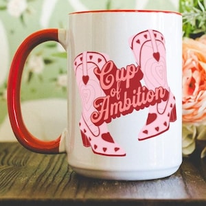 May include: White ceramic mug with a red rim and handle. The mug features a pink and red graphic of two cowboy boots with hearts and the text "Cup of Ambition".