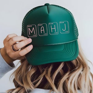Peut inclure: Un chapeau de camionneur vert avec un filet noir à l'arrière. Le chapeau a un motif rose et blanc avec les lettres "MAHJ" dessus.