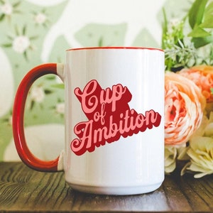 May include: White ceramic mug with a red interior and handle. The mug features the text "Cup of Ambition" in a retro, cursive font. The mug is set on a dark wooden surface with flowers in the background.