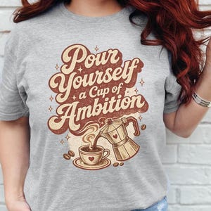 Puede incluir: Una camiseta gris jaspeado con un estampado gr&aacute;fico marr&oacute;n que incluye el texto Pour Yourself a Cup of Ambition. El dise&ntilde;o muestra una cafetera italiana vertiendo caf&eacute; en una taza, rodeada de peque&ntilde;as estrellas y granos de caf&eacute;.