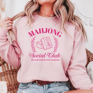 May include: Pink sweatshirt with the text "Mahjong Social Club" and a graphic of two mahjong tiles. The text "Life is better playing Mahjong" is below the graphic.