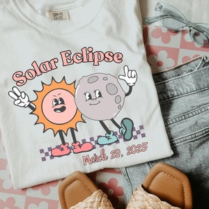 Może przedstawiać: Biała koszulka z grafiką przedstawiającą kreskówkowe słońce i księżyc z tekstem "Solar Eclipse" i "March 29, 2025".