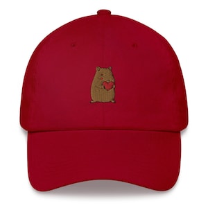 May include: A red baseball cap with an embroidered image of a brown bear holding a red heart.
