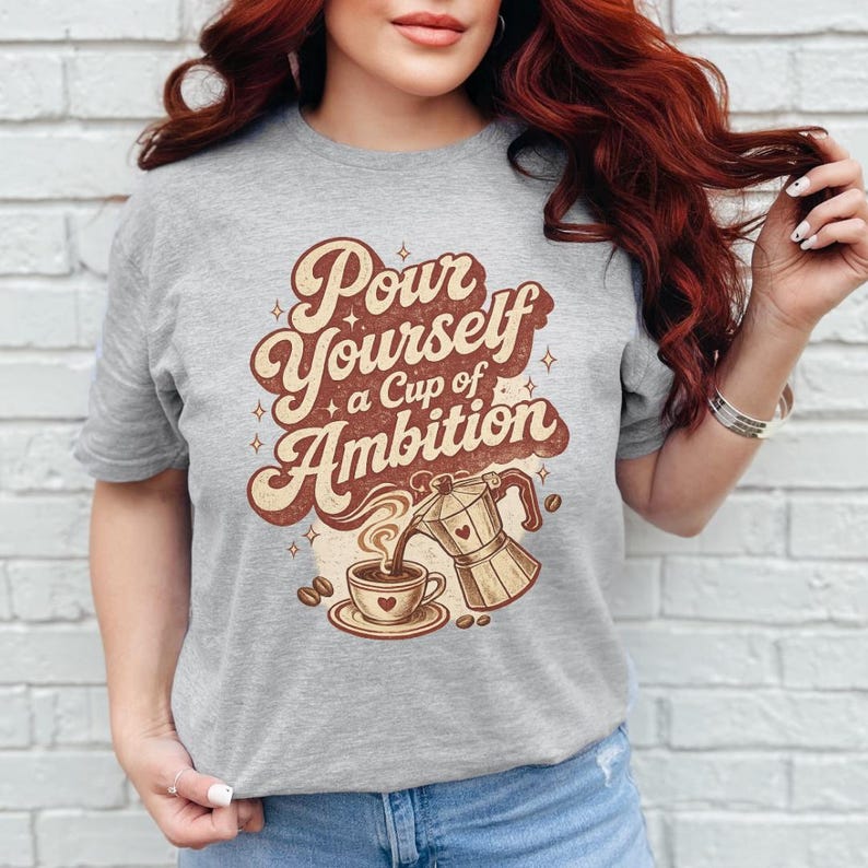 Puede incluir: Una camiseta gris jaspeado con un estampado gr&aacute;fico marr&oacute;n que incluye el texto Pour Yourself a Cup of Ambition. El dise&ntilde;o muestra una cafetera italiana vertiendo caf&eacute; en una taza, rodeada de peque&ntilde;as estrellas y granos de caf&eacute;.