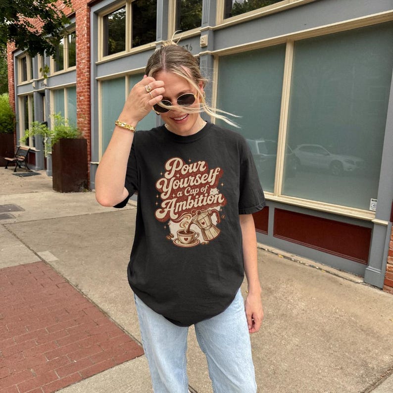 Puede incluir: Una camiseta negra de manga corta con un estampado gr&aacute;fico que incluye el texto Pour Yourself a Cup of Ambition en una fuente retro. El dise&ntilde;o muestra una cafetera italiana vertiendo caf&eacute; en una taza con un coraz&oacute;n y estrellas.