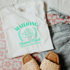 May include: White t-shirt with a green graphic design that says "Mahjong Social Club" and "Life is better playing Mahjong".
