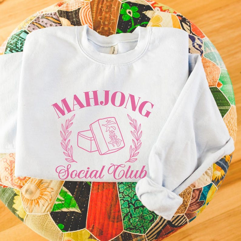 May include: A light blue sweatshirt with the text "Mahjong Social Club" in pink lettering. The design features a stylized image of a mahjong tile.