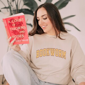 Pode incluir: Uma mulher vestindo uma camiseta bege com a palavra "BOOKWORM" impressa em amarelo na frente. Ela está segurando um livro intitulado "A Court of Thorns and Roses" de Sarah J. Maas.