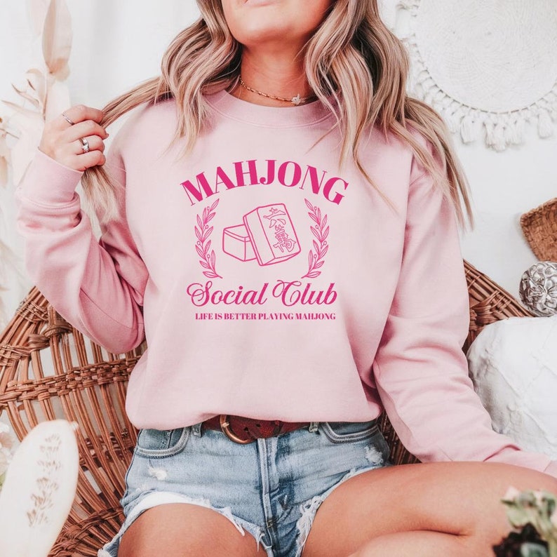 May include: Pink sweatshirt with the text "Mahjong Social Club" and a graphic of two mahjong tiles. The text "Life is better playing Mahjong" is below the graphic.