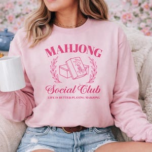 Woman wearing a pink Mahjong Social Club sweatshirt with mahjong tile graphic while relaxing with a coffee mug, cozy gift for mahjong lovers and game night players.