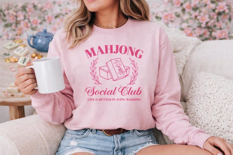 Woman wearing a pink Mahjong Social Club sweatshirt with mahjong tile graphic while relaxing with a coffee mug, cozy gift for mahjong lovers and game night players.