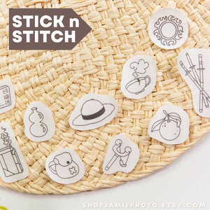 May include: A set of ten black and white iron-on patches featuring various designs, including a book, a gourd, a hat, a cup of tea, a slingshot, a backpack, a citrus fruit, a sun, swords, and a cigarette pack. The patches are on a woven straw mat with the text "STICK n STITCH" above them.