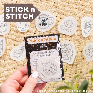 May include: A set of iron-on patches featuring various illustrations, including a cat in a mug, a leaf, a spiral, a Totoro character, a No Face character, a Spirited Away character, and a guitar. The patches are on a woven straw background. The text "Stick n' Stitch" and "Your order means the world" is on a black background with gold stars.
