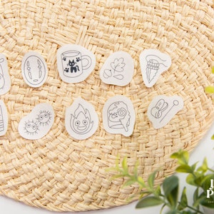 May include: A set of 10 black and white illustrated stickers featuring characters and objects from the Studio Ghibli film, Spirited Away. The stickers are on a woven straw background.
