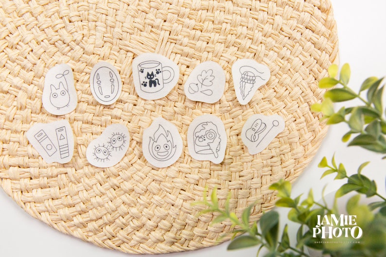 May include: A set of 10 black and white illustrated stickers featuring characters and objects from the Studio Ghibli film, Spirited Away. The stickers are on a woven straw background.