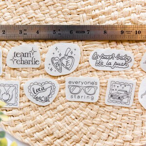 May include: A collection of ten white adhesive stickers with black line art designs, including a cactus, a locket, a wolf, a truck, and text such as team charlie, la push baby, and cullen, displayed with a 12-inch stainless steel ruler.