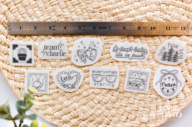 May include: A collection of ten white adhesive stickers with black line art designs, including a cactus, a locket, a wolf, a truck, and text such as team charlie, la push baby, and cullen, displayed with a 12-inch stainless steel ruler.