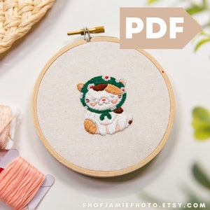 May include: A white cat with brown and orange markings wearing a green and white floral hooded hat. The cat is embroidered on a white fabric stretched over a wooden embroidery hoop. The hoop is on a white surface.