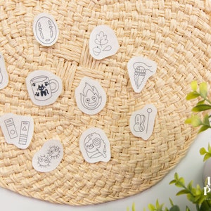 May include: A set of 10 white stickers with black line drawings of various characters and objects, including a cat, a cup of coffee, a fire spirit, a Totoro, a book, a needle and thread, a leaf, a cat, and a cat with a hat. The stickers are arranged on a woven straw mat.