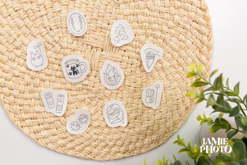 May include: A set of 10 white stickers with black line drawings of various characters and objects, including a cat, a cup of coffee, a fire spirit, a Totoro, a book, a needle and thread, a leaf, a cat, and a cat with a hat. The stickers are arranged on a woven straw mat.
