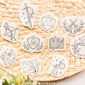 May include: A collection of ten white stickers featuring black line art illustrations of fantasy items including a sword, a twenty-sided die, a bow, a book, a cauldron, a map, a key, a crystal, a potion bottle, and a compass.