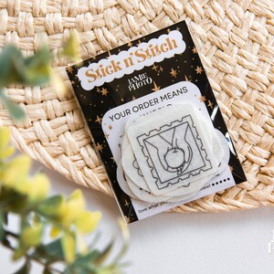 May include: A stack of white Stick n' Stitch embroidery stabilizer sheets featuring a black line art design of hands holding an apple inside a postage stamp border, packaged on a black card with gold stars and the text Stick n' Stitch.
