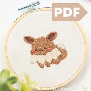 以下が含まれることがあります： 白い布地に刺繍された茶色とクリーム色のポケモンイーブイ。木製刺繍枠に入っています。枠は白い背景の上に置かれています。