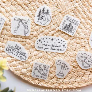 May include: A collection of ten white fabric patches featuring black line art designs including a wolf, a bow with the word forever, a baseball cap, a stamp labeled Forks, a truck, a chess piece, and text quotes from Twilight.