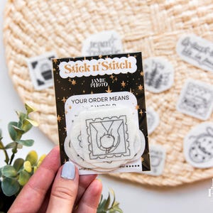 May include: A black and white Stick n'Stitch embroidery stabilizer pack featuring a central square patch with a line art illustration of hands holding an apple. The packaging includes gold star accents and the text Jamie Photo.
