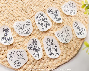 Op de afbeelding: Een verzameling van tien witte, onregelmatig gevormde stickers met zwarte lijntekeningen van diverse bloemstengels, bladeren en kleine stermotieven. De stickers zijn gerangschikt op een geweven, lichtbruin, cirkelvormig oppervlak.