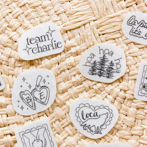 May include: A collection of white fabric patches featuring black line art designs including a cactus, a heart locket with the letters B and E, a wolf inside a heart, a truck, pine trees, and text reading team charlie and la push baby.