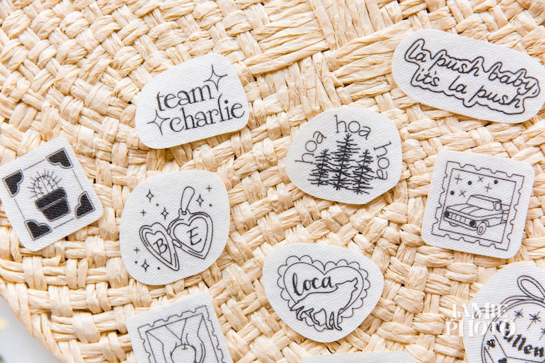 May include: A collection of white fabric patches featuring black line art designs including a cactus, a heart locket with the letters B and E, a wolf inside a heart, a truck, pine trees, and text reading team charlie and la push baby.