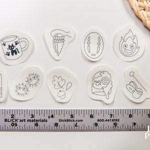 May include: Ten white fabric patches with black ink drawings of characters and objects from the animated film 'My Neighbor Totoro'. The patches include a cat, a cup, a leaf, a spirit, a bus, a soot sprite, a mushroom, a Totoro, and a No Face.