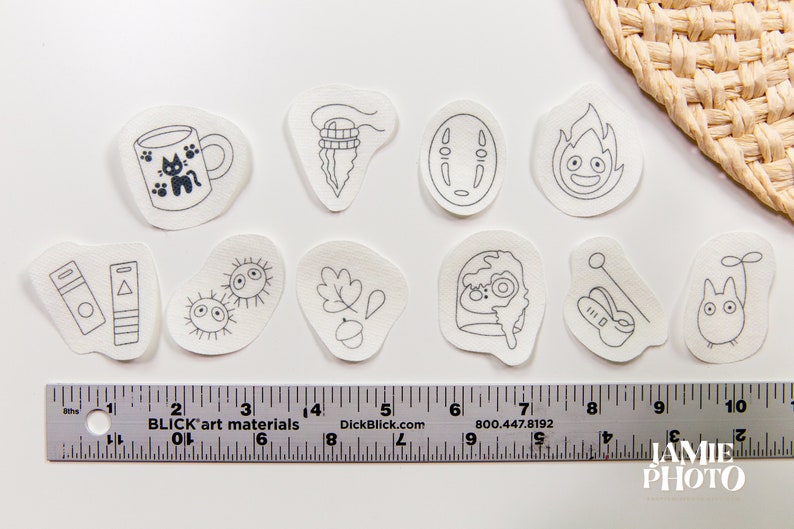 May include: Ten white fabric patches with black ink drawings of characters and objects from the animated film 'My Neighbor Totoro'. The patches include a cat, a cup, a leaf, a spirit, a bus, a soot sprite, a mushroom, a Totoro, and a No Face.