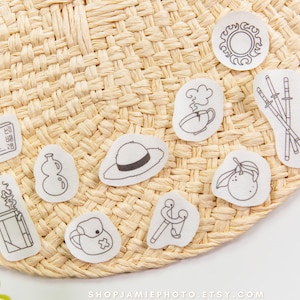 Puede incluir: Una colección de nueve pegatinas blancas individuales con ilustraciones de líneas negras de varios objetos, incluyendo un sol, una taza, espadas, un libro, una botella, un sombrero, una fruta, un encendedor y una mochila.